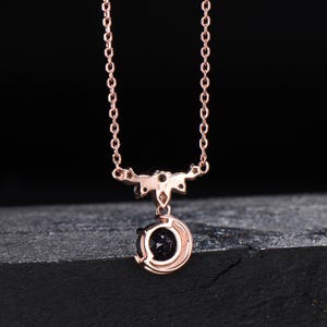 May include: A rose gold necklace featuring a delicate chain and a pendant shaped like a crescent moon. The pendant holds a round, dark blue, glittery stone at its center. The metal setting includes small star-shaped accents above the moon.
