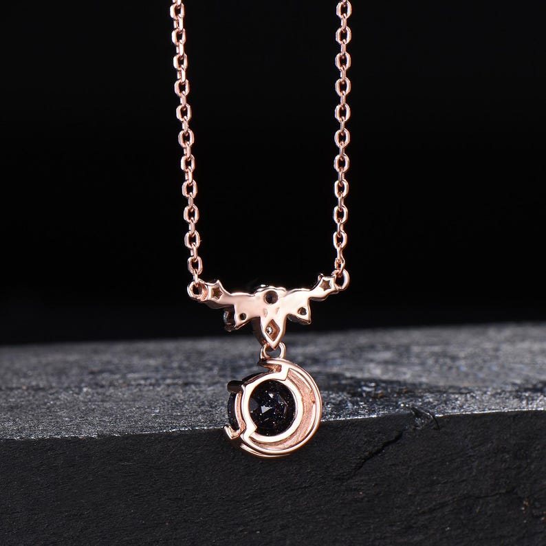 May include: A rose gold necklace featuring a delicate chain and a pendant shaped like a crescent moon. The pendant holds a round, dark blue, glittery stone at its center. The metal setting includes small star-shaped accents above the moon.