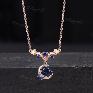 May include: A rose gold necklace featuring a delicate chain and a celestial-themed pendant. The pendant displays a round, dark blue glittery stone set within a crescent moon, accented by small blue and clear stones, stars, and the text DRingstudio.