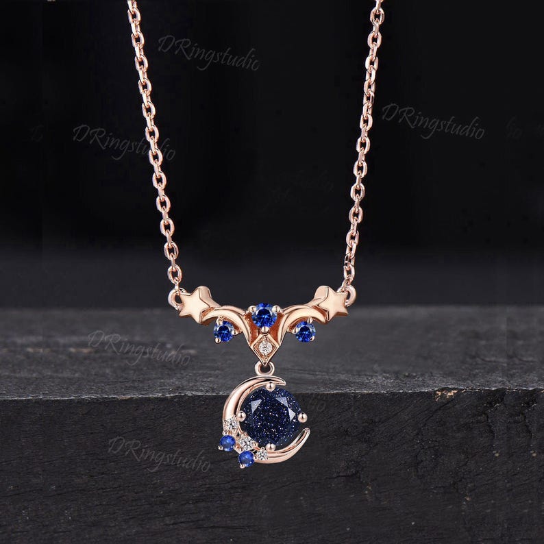 May include: A rose gold necklace featuring a delicate chain and a celestial-themed pendant. The pendant displays a round, dark blue glittery stone set within a crescent moon, accented by small blue and clear stones, stars, and the text DRingstudio.