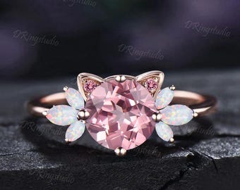 May include: A rose gold ring featuring a large pink gemstone center, cat ear accents, and white opal flower-shaped side stones. The ring has a delicate, feminine design, perfect for a unique jewelry piece.