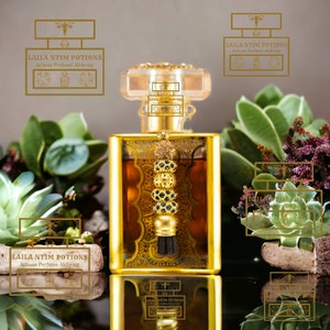 May include: A gold and clear glass bottle of perfume with a gold tassel and a black and gold stopper. The bottle is sitting on a black surface with a reflection of the bottle. The bottle is surrounded by green succulents and the text "LAILA NTIM POTIONS Artisan Perfume Alchemy" is visible on the bottle and in the reflection.