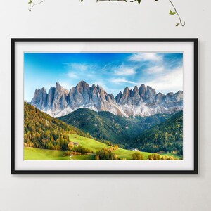 Puede incluir: Una impresión enmarcada de un paisaje de montañas bajo un cielo azul con nubes. Las montañas son grises y blancas, con árboles verdes y amarillos en primer plano. Se ve un valle verde y pequeños edificios.