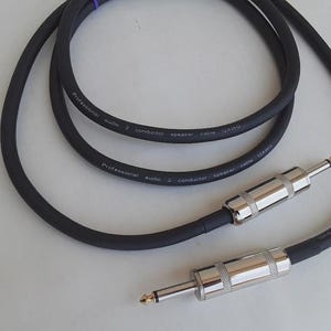 May include: Black audio speaker cable with two 1/4 inch (6.35 mm) male connectors. The cable is labeled "Professional audio 2 conductor speaker cable 12AWG". The connectors have silver metal housings and gold-colored tips.