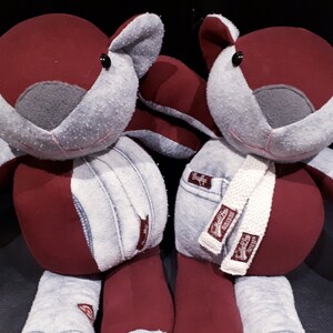 May include: Two plush squirrel toys made from recycled denim and fabric. The squirrels are burgundy and grey with black eyes and are sitting on a dark surface.