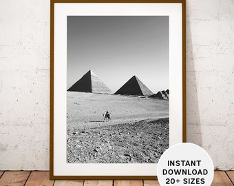 Könnte beinhalten: Schwarzweißfoto der drei großen Pyramiden von Gizeh in Ägypten. Im Vordergrund geht ein Kamel mit seinem Reiter durch die Wüste.
