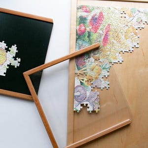 May include: A wooden-framed puzzle board with a partially assembled jigsaw puzzle. The puzzle features a colorful floral design. A separate wooden frame with a clear acrylic sheet is also present.