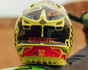 May include: A lime green and black motocross helmet with the word "GUDAC" in orange lettering. The helmet is resting on a green motorcycle. The exhaust pipe is visible.