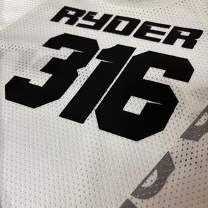 K&ouml;nnte beinhalten: Wei&szlig;es Trikot mit schwarzem Netzstoff. Das Trikot hat die schwarze Nummer "316" und den Namen "Ryder" auf der R&uuml;ckseite.