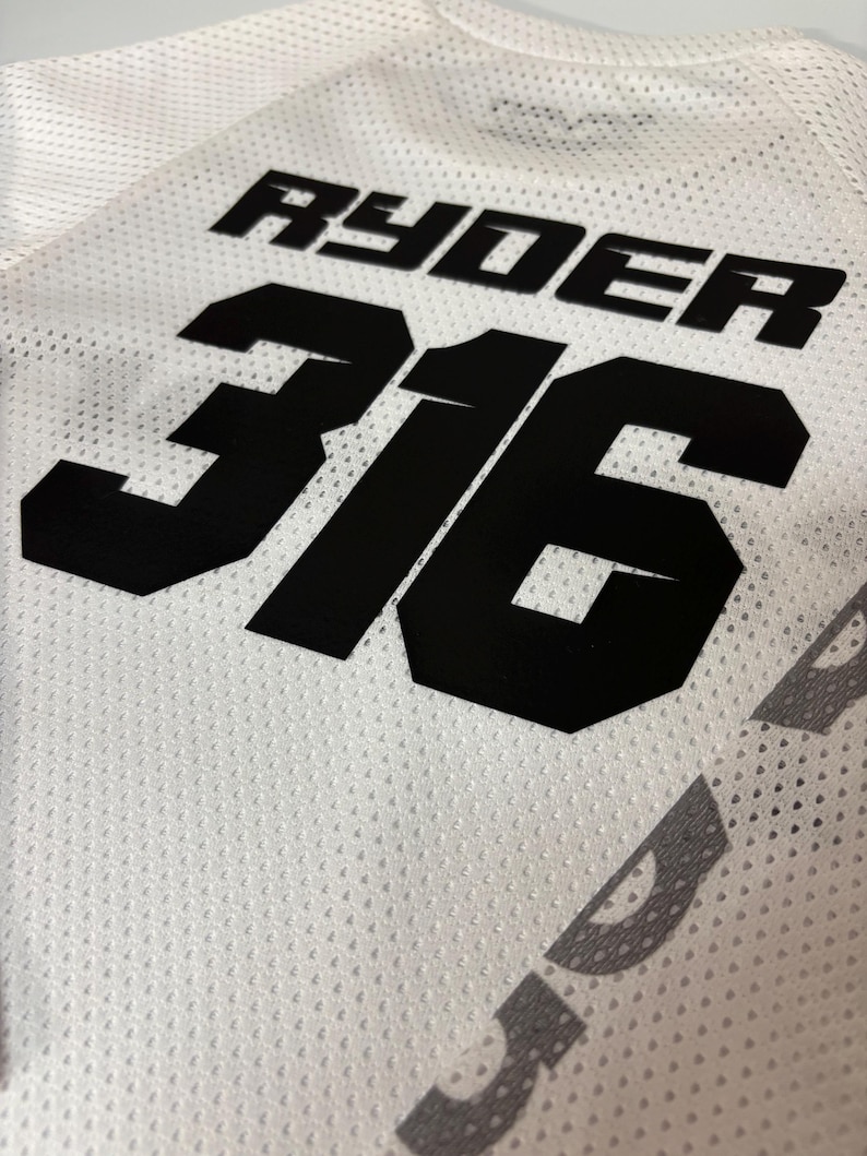 K&ouml;nnte beinhalten: Wei&szlig;es Trikot mit schwarzem Netzstoff. Das Trikot hat die schwarze Nummer "316" und den Namen "Ryder" auf der R&uuml;ckseite.