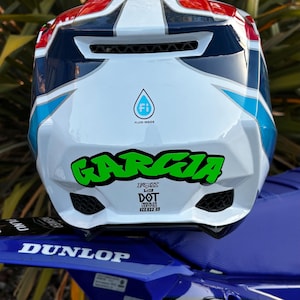 Könnte beinhalten: Ein weißer, blauer und roter Motocross-Helm mit einem Fluid Inside-Logo, einem grünen Garcia-Aufkleber sowie DOT- und ECE 22-05-Zertifizierungsetiketten. Der Helm liegt auf einem blauen Motorradschutzblech mit weißem Dunlop-Logo und Motocutz-Text.