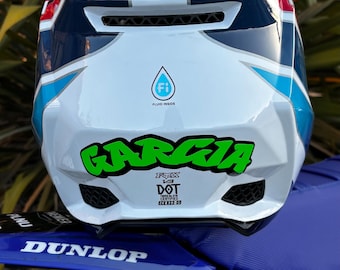 Può includere: Un casco da motocross bianco, blu e rosso con logo Fluid Inside, adesivo verde Garcia ed etichette di certificazione DOT ed ECE 22-05. Il casco è posizionato su un parafango blu con logo Dunlop bianco e la scritta Motocutz.