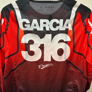 K&ouml;nnte beinhalten: Rotes, schwarzes und wei&szlig;es Motocross-Trikot mit dem Namen "Garcia" und der Nummer "316" auf der R&uuml;ckseite. Das Trikot tr&auml;gt auch den Schriftzug "Premier" auf der R&uuml;ckseite.