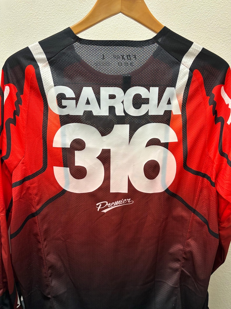 K&ouml;nnte beinhalten: Rotes, schwarzes und wei&szlig;es Motocross-Trikot mit dem Namen "Garcia" und der Nummer "316" auf der R&uuml;ckseite. Das Trikot tr&auml;gt auch den Schriftzug "Premier" auf der R&uuml;ckseite.
