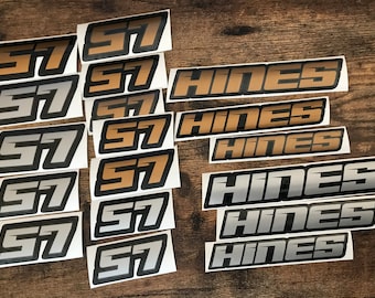 Può includere: Una collezione di adesivi con il numero "57" e il nome "HINES" in varie combinazioni di colori. Gli adesivi hanno un contorno nero con riempimenti dorati, argentati e beige. Gli adesivi sono su una superficie di legno.