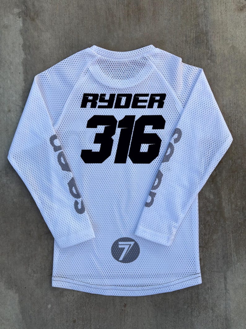 K&ouml;nnte beinhalten: Wei&szlig;es Langarm-Trikot mit schwarzem Netzstoff. Auf der R&uuml;ckseite des Trikots steht der Name "RYDER" und die Nummer "316" in Schwarz. Die &Auml;rmel haben ein graues "7"-Logo.
