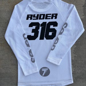 K&ouml;nnte beinhalten: Wei&szlig;es Langarm-Trikot mit schwarzem Netzstoff. Auf der R&uuml;ckseite des Trikots sind der Name "Ryder" und die Nummer "316" in Schwarz gedruckt. Der linke &Auml;rmel hat einen grauen Kreis mit der Nummer "7" in Schwarz gedruckt.