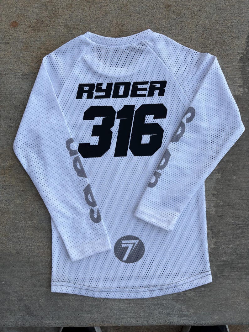 K&ouml;nnte beinhalten: Wei&szlig;es Langarm-Trikot mit schwarzem Netzstoff. Auf der R&uuml;ckseite des Trikots sind der Name "Ryder" und die Nummer "316" in Schwarz gedruckt. Der linke &Auml;rmel hat einen grauen Kreis mit der Nummer "7" in Schwarz gedruckt.