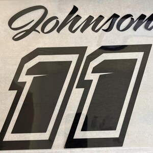 K&ouml;nnte beinhalten: Schwarzer Vinyl-Aufkleber mit dem Namen "Johnson" und der Nummer "11" in einer fetten, umrandeten Schrift.