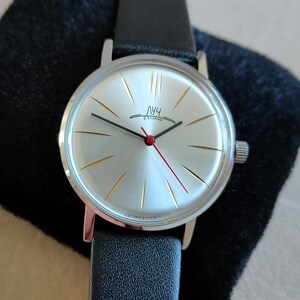 May include: A silver wristwatch with a white face and black leather band. The watch face has gold-colored markings and a red second hand. The watch face also has the text "Луч" printed on it.