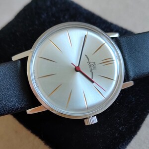 May include: A silver wristwatch with a white face and black leather band. The watch face has gold-colored hour markers and a red second hand. The watch face also has the text "HWU 23 Jewels" printed on it.