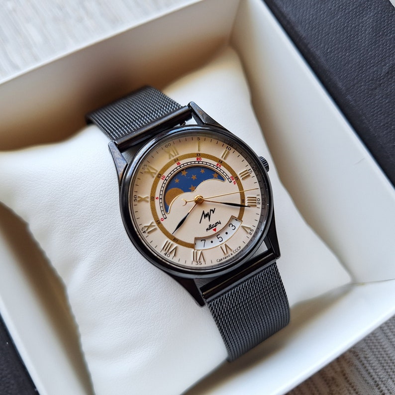 May include: A black wristwatch with a silver face and a black mesh band. The face has Roman numeral hour markers, a moon phase indicator, and the text "Чайка СССР".