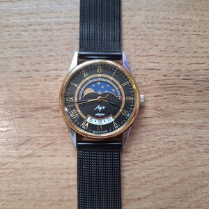 May include: A vintage wristwatch with a black face, gold-tone bezel, and a black mesh band. The watch face features Roman numerals, a moon phase indicator, and the Cyrillic word "Лучь". The date is displayed at the bottom. The watch is on a wooden surface.