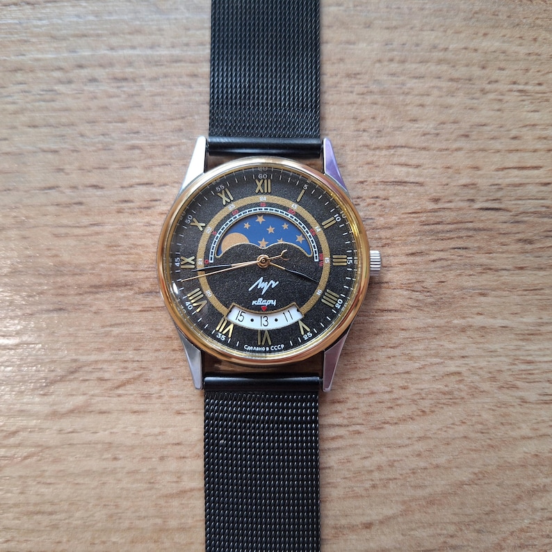May include: A vintage wristwatch with a black face, gold-tone bezel, and a black mesh band. The watch face features Roman numerals, a moon phase indicator, and the Cyrillic word "Лучь". The date is displayed at the bottom. The watch is on a wooden surface.