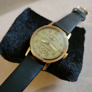 Puede incluir: Un reloj de pulsera dorado con una correa de cuero negro. La esfera del reloj es de color crema con n&uacute;meros negros y una aguja de segundos roja. La esfera del reloj tambi&eacute;n presenta el texto "ТУРМАНСКНЕ" y "ПЕРВЫЙ ЧЕЛОВЕК В КОСМОС" con una peque&ntilde;a imagen de un cohete. El texto "12 АПРЕЛЯ 1961" y "ЮРИЙ ГАГАРИН" tambi&eacute;n est&aacute; en la esfera del reloj.