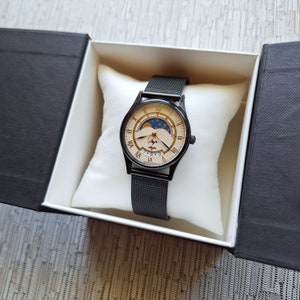 May include: A black watch with a white face and gold accents. The watch has a moon phase complication and a black mesh band. The watch is in a white box with a black lid.