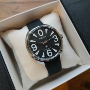 Puede incluir: Un reloj analógico negro con números y manecillas blancas, con el nombre de la marca "PAKETA" en la esfera. El reloj tiene una correa negra y un bisel plateado. El reloj se presenta en una caja blanca con exterior negro.