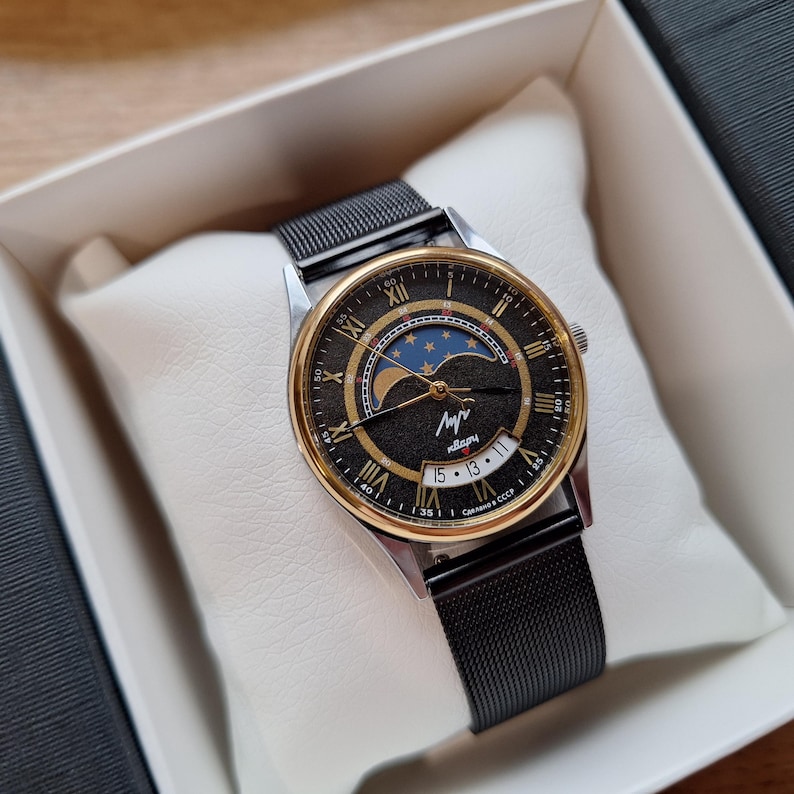May include: A vintage wristwatch with a black mesh band and a gold-tone bezel. The watch face features Roman numerals, a moon phase indicator, and the text "Made in USSR". The watch is presented in a white box.