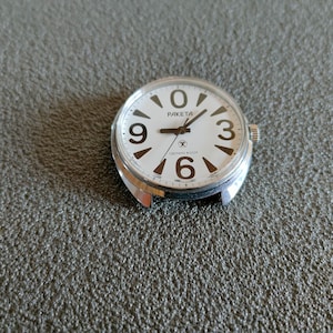 May include: A vintage Raketa watch face with a white dial and large black numerals. The watch face has the brand name "RAKETA" and Cyrillic text. The watch has a silver-coloured metal casing.