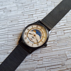 May include: A black wristwatch with a white face and gold Roman numeral hour markers. The watch has a moon phase complication and a date window. The watch is on a black metal mesh band.