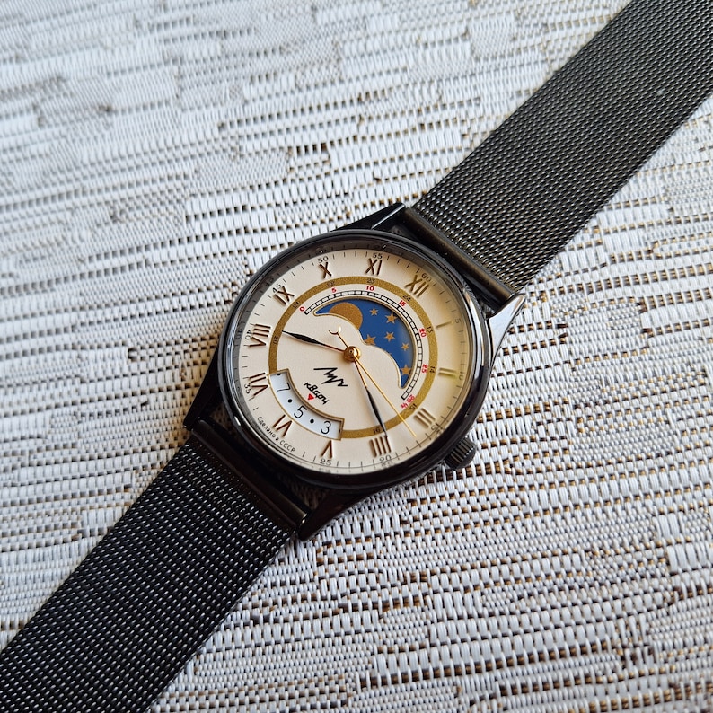 May include: A black wristwatch with a white face and gold Roman numeral hour markers. The watch has a moon phase complication and a date window. The watch is on a black metal mesh band.