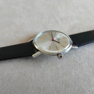 May include: A silver wristwatch with a white face and black leather band. The watch face has gold-colored markings and a red second hand. The watch face also has the text "INU" in black.