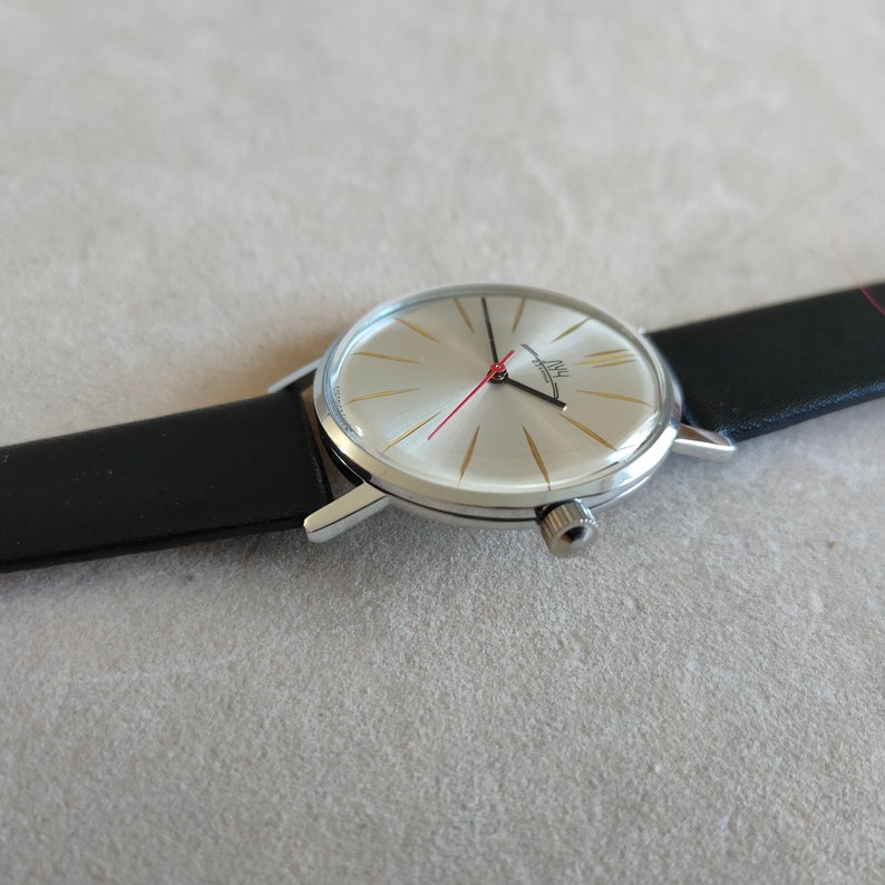 May include: A silver wristwatch with a white face and black leather band. The watch face has gold-colored markings and a red second hand. The watch face also has the text "INU" in black.