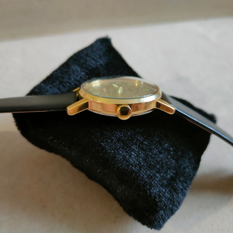 Puede incluir: Un reloj de pulsera de tono dorado con una correa de cuero negro. La esfera del reloj tiene un fondo blanco con marcas negras y una aguja de segundos de tono dorado.
