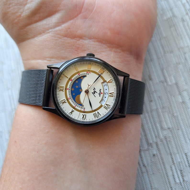 May include: A black wristwatch with a white face and gold Roman numeral hour markers. The watch has a moon phase complication and a date window. The watch is on a black metal mesh band. The watch face says "Мир" and "Made in USSR".