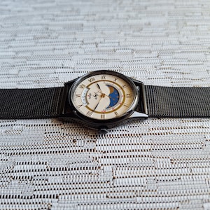 May include: A black wristwatch with a white face and gold accents. The watch has a moon phase complication and a date window. The watch is on a black metal band.
