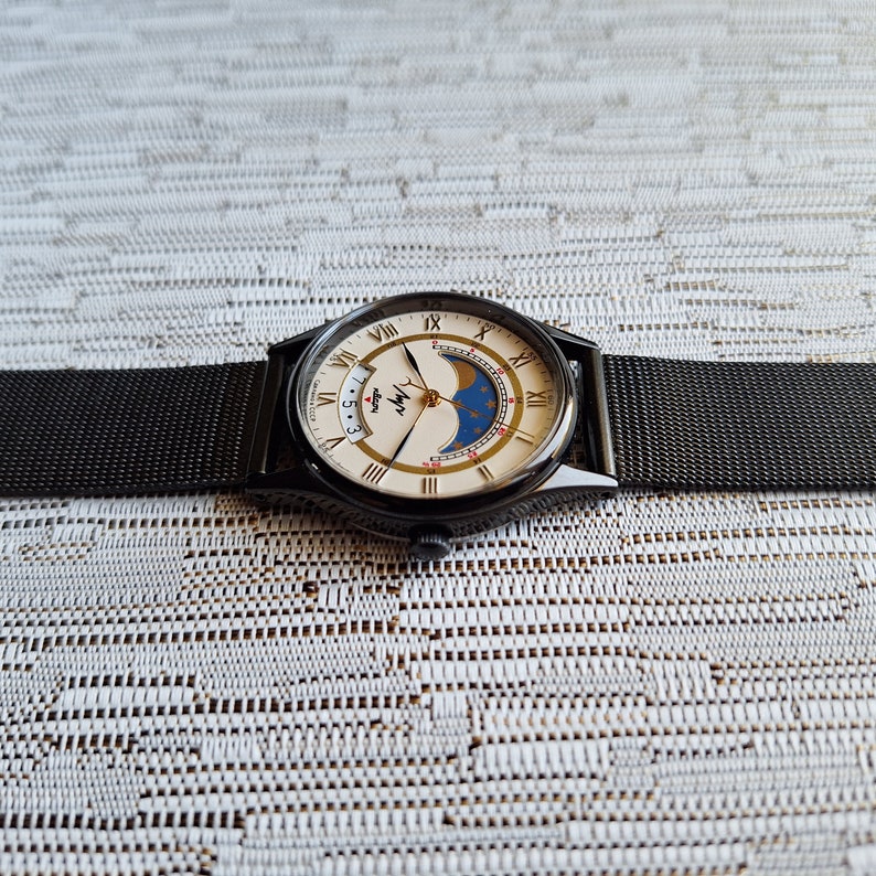 May include: A black wristwatch with a white face and gold accents. The watch has a moon phase complication and a date window. The watch is on a black metal band.