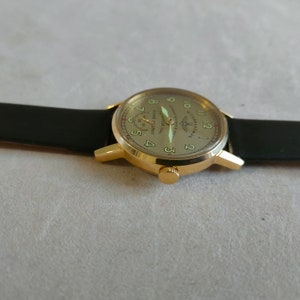 Puede incluir: Un reloj de pulsera dorado con una correa de cuero negro. La esfera del reloj es de color crema con marcadores de hora en n&uacute;meros romanos negros y marcadores de minutos negros. La esfera del reloj tambi&eacute;n tiene un peque&ntilde;o dial de segundos en la posici&oacute;n de las 9 en punto y el texto "1961 год. Первый полёт человека в космос" en escritura cir&iacute;lica.