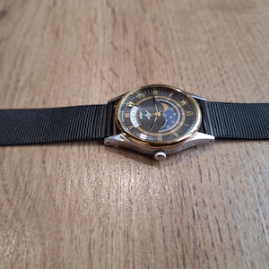 May include: A wristwatch with a black mesh band and a silver and gold-toned case. The watch face is dark with gold Roman numerals and a moon phase indicator. The watch is displayed on a wooden surface.