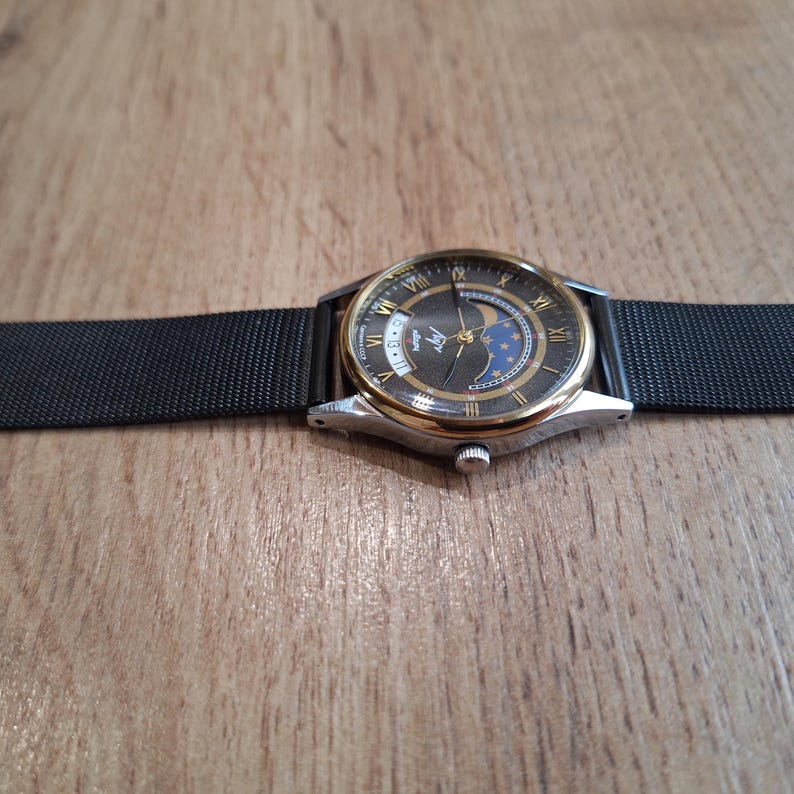 May include: A wristwatch with a black mesh band and a silver and gold-toned case. The watch face is dark with gold Roman numerals and a moon phase indicator. The watch is displayed on a wooden surface.