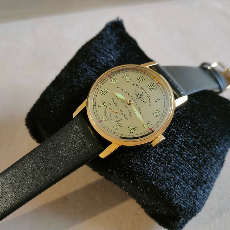 Puede incluir: Un reloj de pulsera dorado con una correa de cuero negro. La esfera del reloj tiene un fondo de color crema con marcadores de hora romanos negros y marcadores de minutos negros. La esfera del reloj tambi&eacute;n presenta una inscripci&oacute;n roja que dice "ПЕРВЫЙ ЧЕЛОВЕК В КОСМОС 12 АПРЕЛЯ 1961 ЮРИЙ ГАГАРИН".