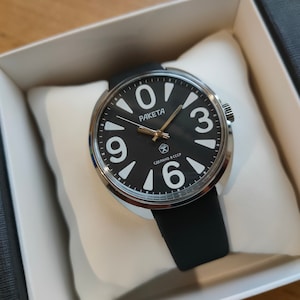 Op de afbeelding: Een zwarte Raketa-horloge met grote witte cijfers en wijzers. Het horloge heeft een zilverkleurige bezel en een zwarte band. Het horloge wordt gepresenteerd in een witte doos met de tekst "СДЕЛАНО В СССР".