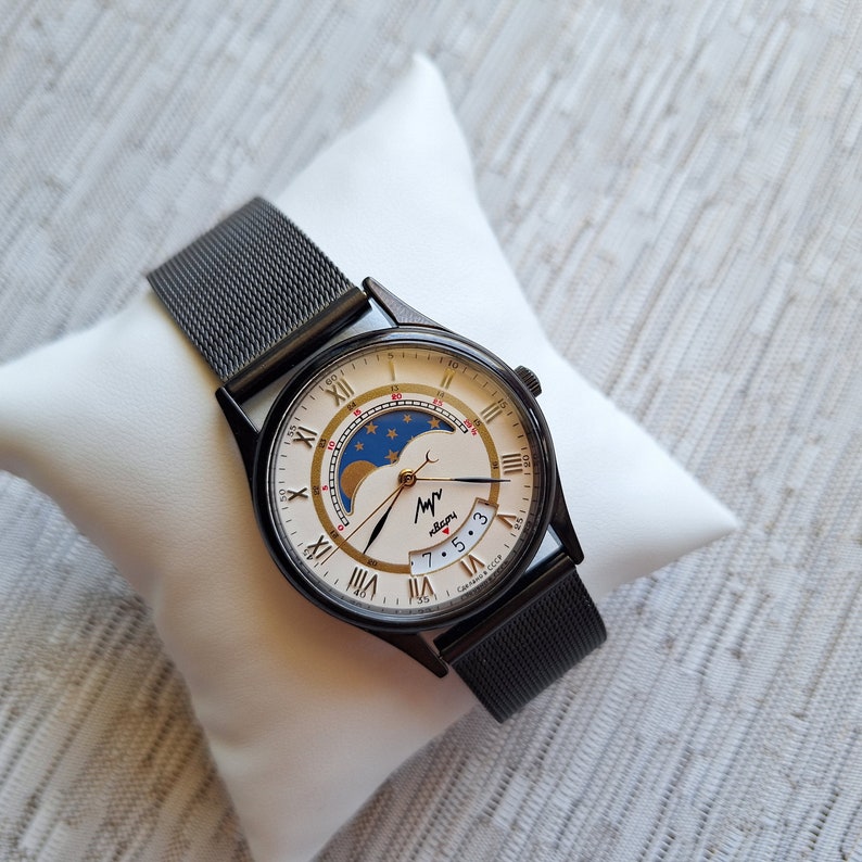 May include: A black watch with a silver face and a black mesh band. The face has Roman numerals and a moon phase indicator. The watch has the text "My Moon" on the face.