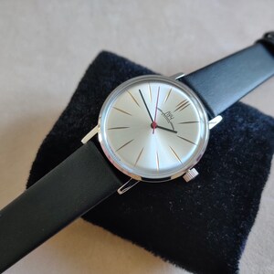 May include: A silver wristwatch with a white face and black leather band. The watch face has gold-colored markings and the text "AWU  EXTON" in black.