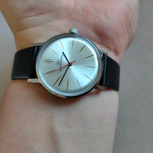 May include: A silver wristwatch with a white face and black hands. The watch has a black leather band and gold accents on the face. The watch face has the word "PAUL" written on it.