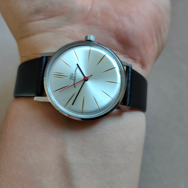 May include: A silver wristwatch with a white face and black hands. The watch has a black leather band and gold accents on the face. The watch face has the word "PAUL" written on it.